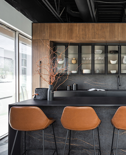 Modern kitchen showroom featuring a sleek dark countertop and island with a natural stone texture, paired with warm brown leather barstools. The backdrop showcases open shelving with glass-front cabinets displaying elegant dishware, framed by vertical wooden slats. Large windows allow natural light to fill the space, highlighting the contemporary and sophisticated design.